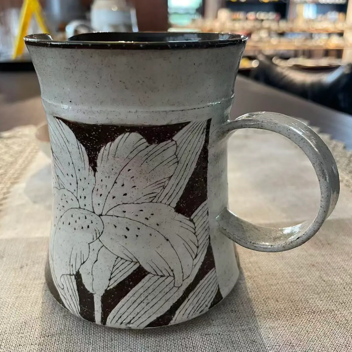 A speckled stoneware creamer with a pinched pouring spout and round handle, white blossoms carved through the dark clay