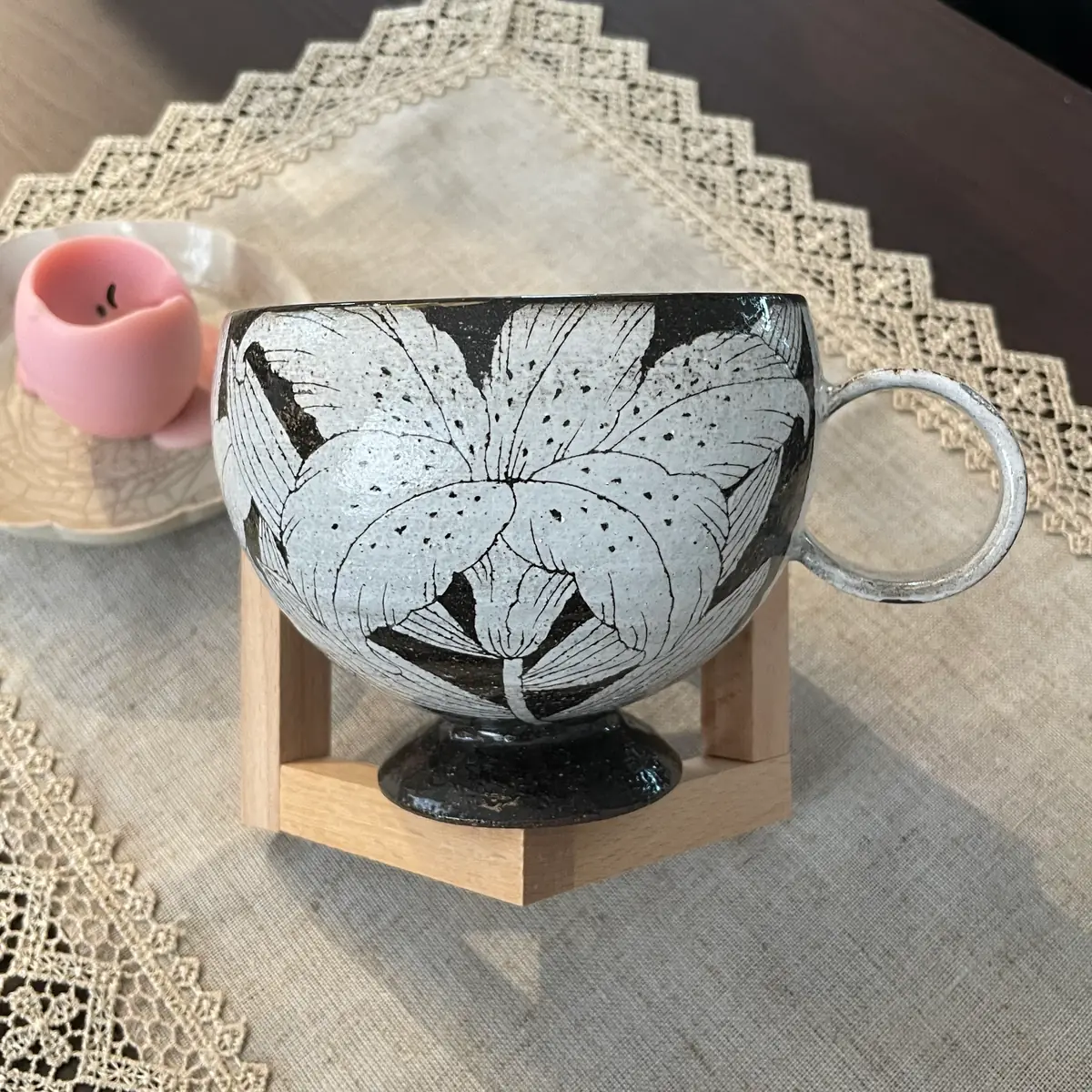 A footed stoneware cup on a wooden stand, pale blossoms carved all round the bowl, a round loop handle at the right and a dark glazed pedestal foot