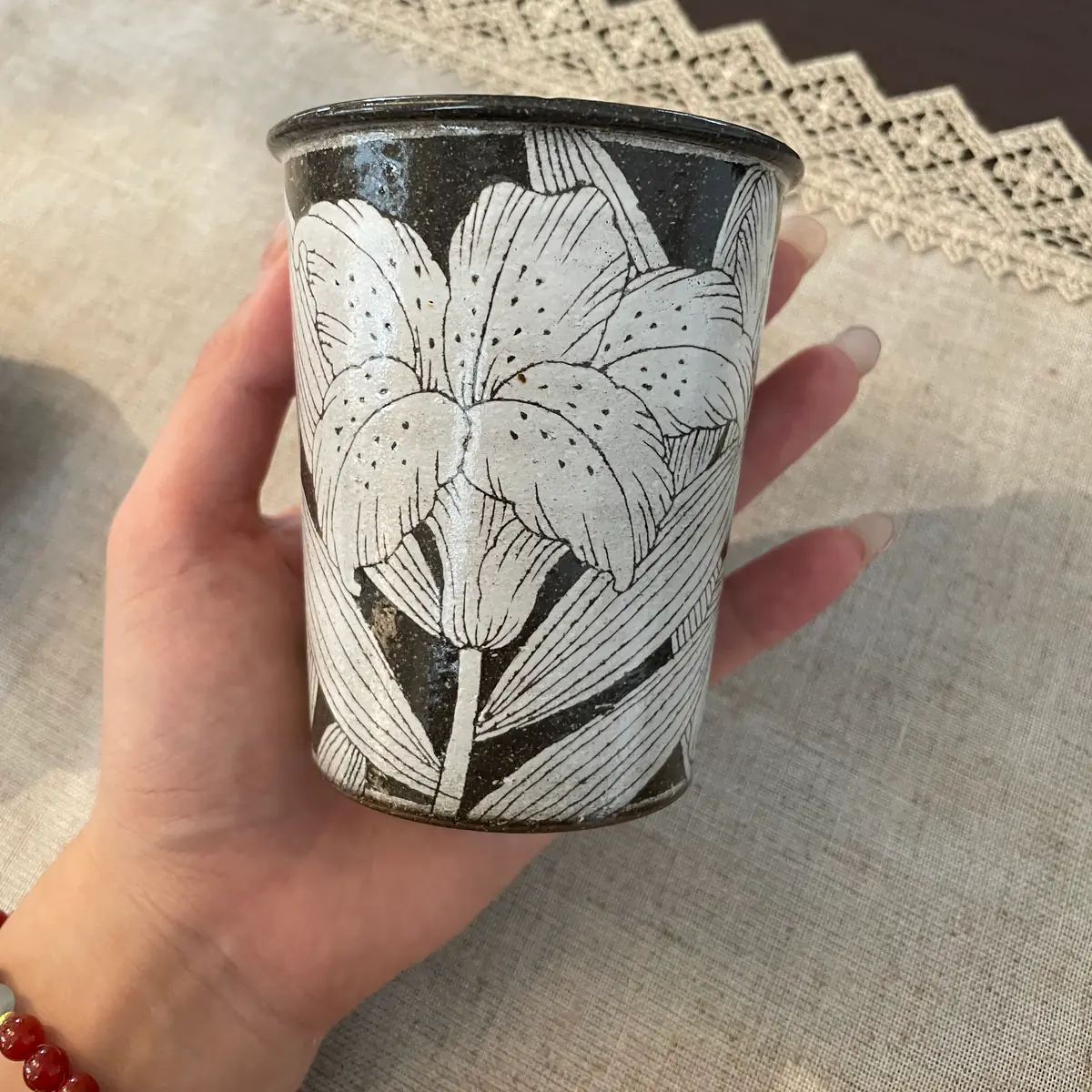 A straight-sided stoneware tumbler held in one hand, pale blossoms flecked with dark specks carved all round the dark clay