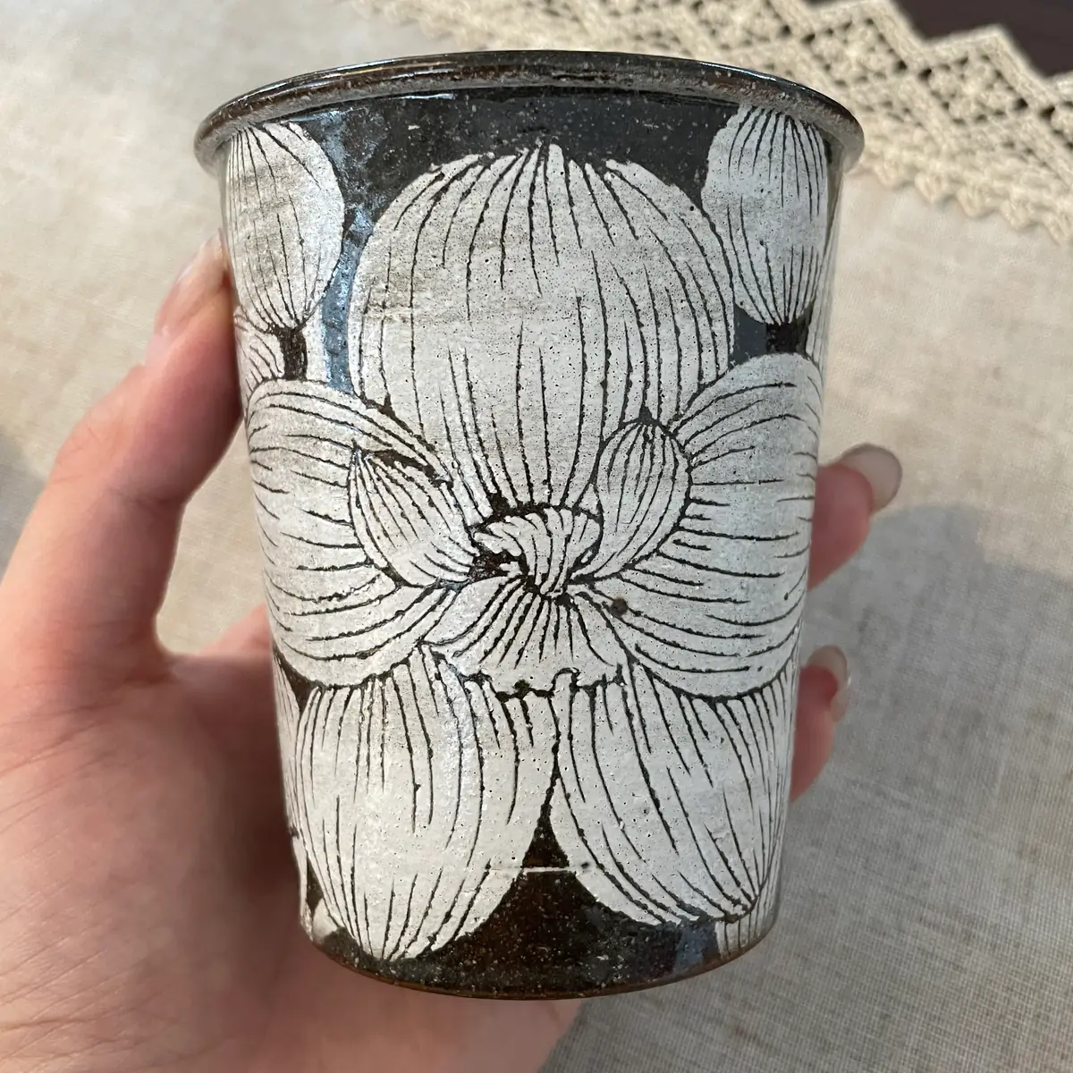 A straight-sided stoneware tumbler held in one hand, one broad blossom carved around the wall in fine parallel lines
