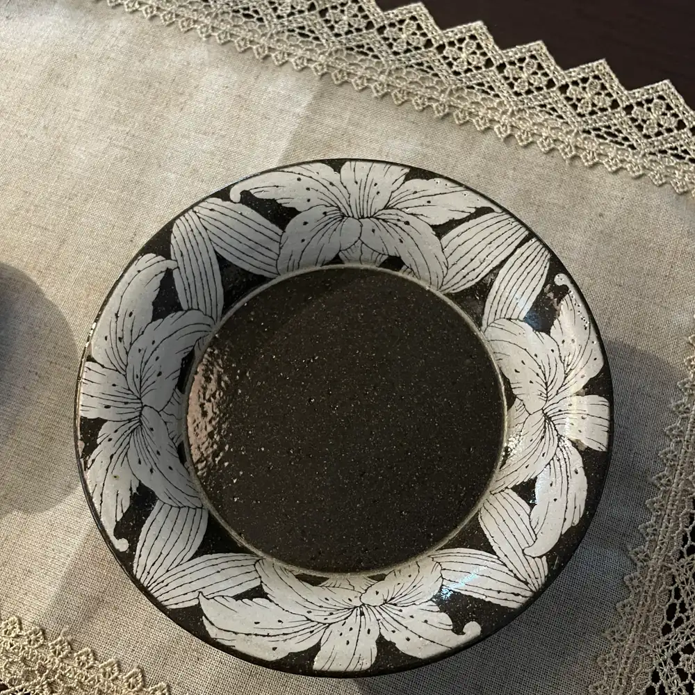 A shallow stoneware dish seen from above, its wide rim carved with white lilies around a dark glazed well