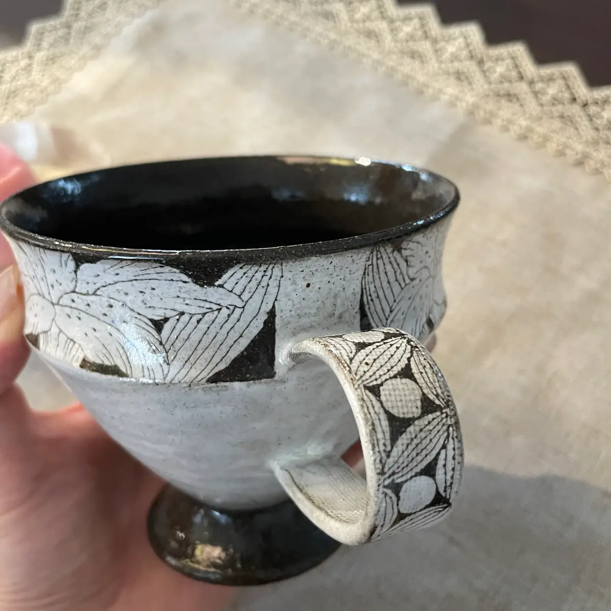 A footed stoneware cup held in one hand, its wide flat strap handle carved with leaves and round fruits, dark glazed interior
