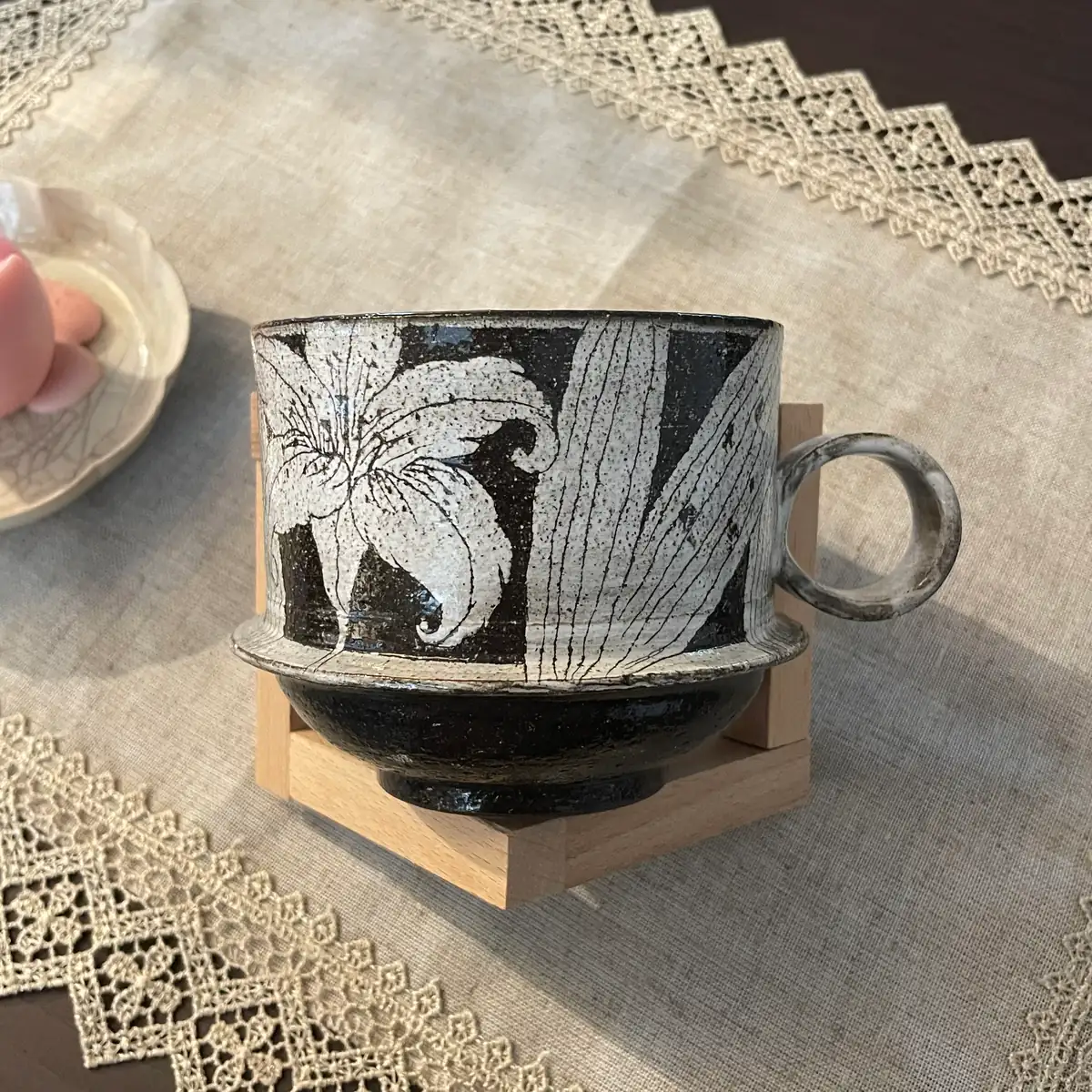 A wide low stoneware cup on a wooden stand, large carved lilies above a stepped flange, dark glazed lower body