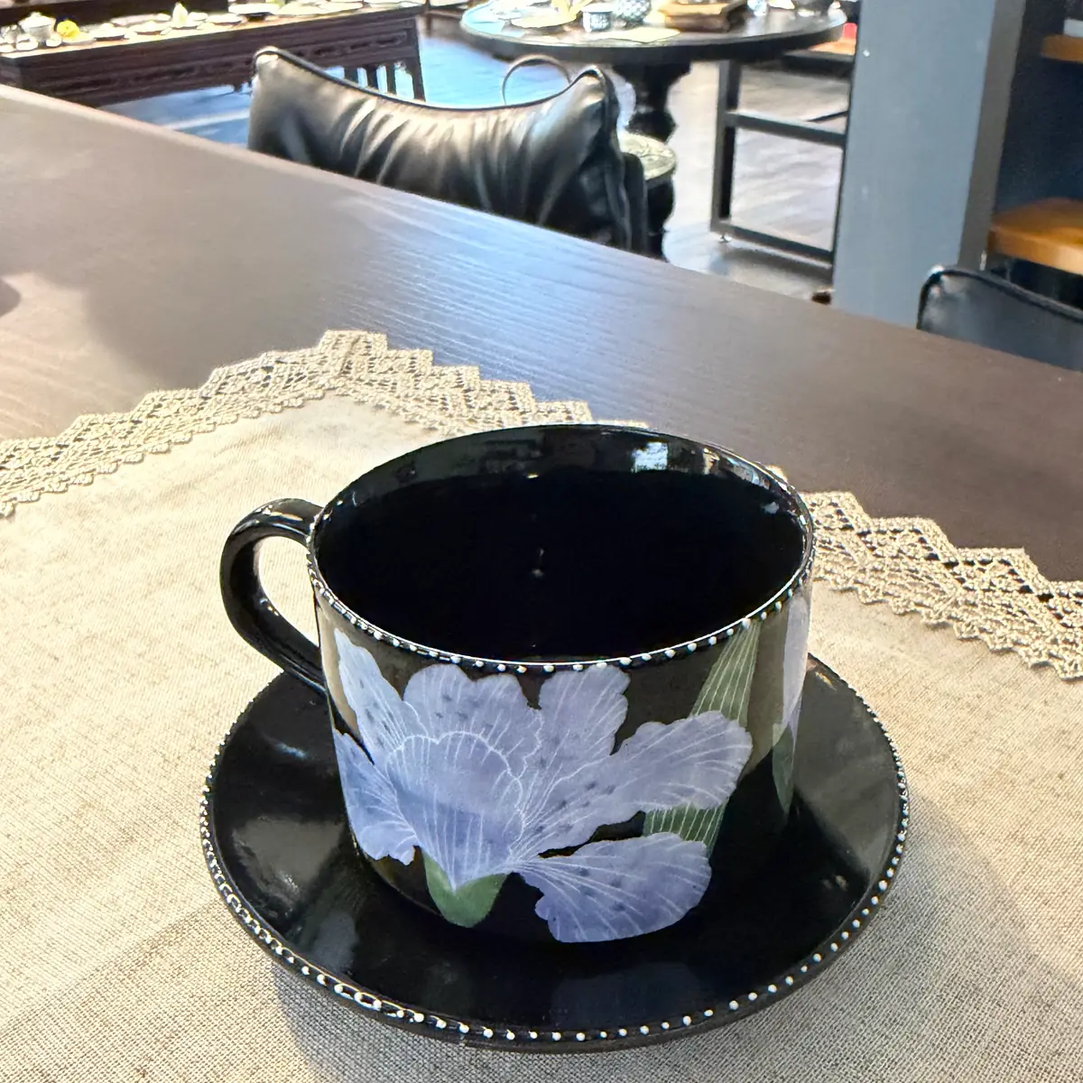 A glossy black cup and saucer painted with pale lilac blossoms and green leaves, both edged with a fine line of white dots