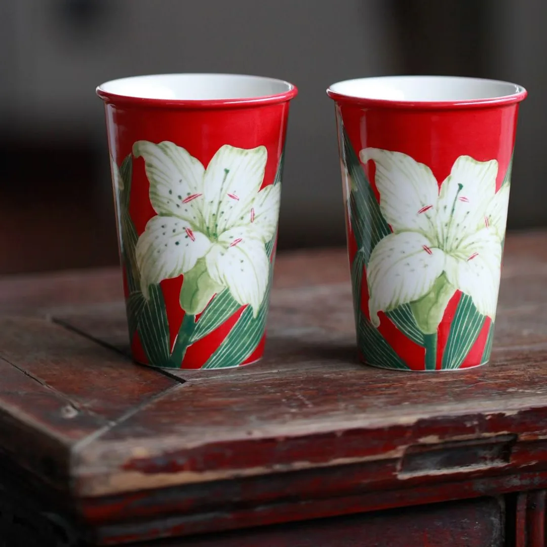 Two tall red-glazed tumblers side by side on an old wooden table, each painted with a large white lily and long green leaves